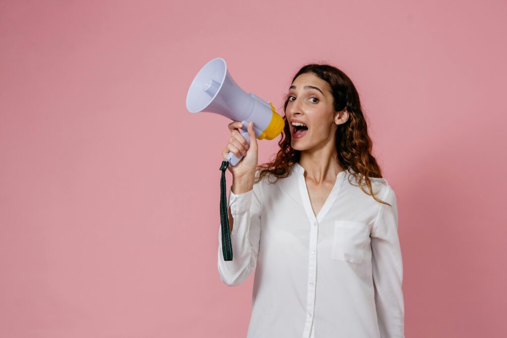 pexels-photo-8638300-8638300 Woman in white shirt holding megaphone against a pink background, confidently speaking.